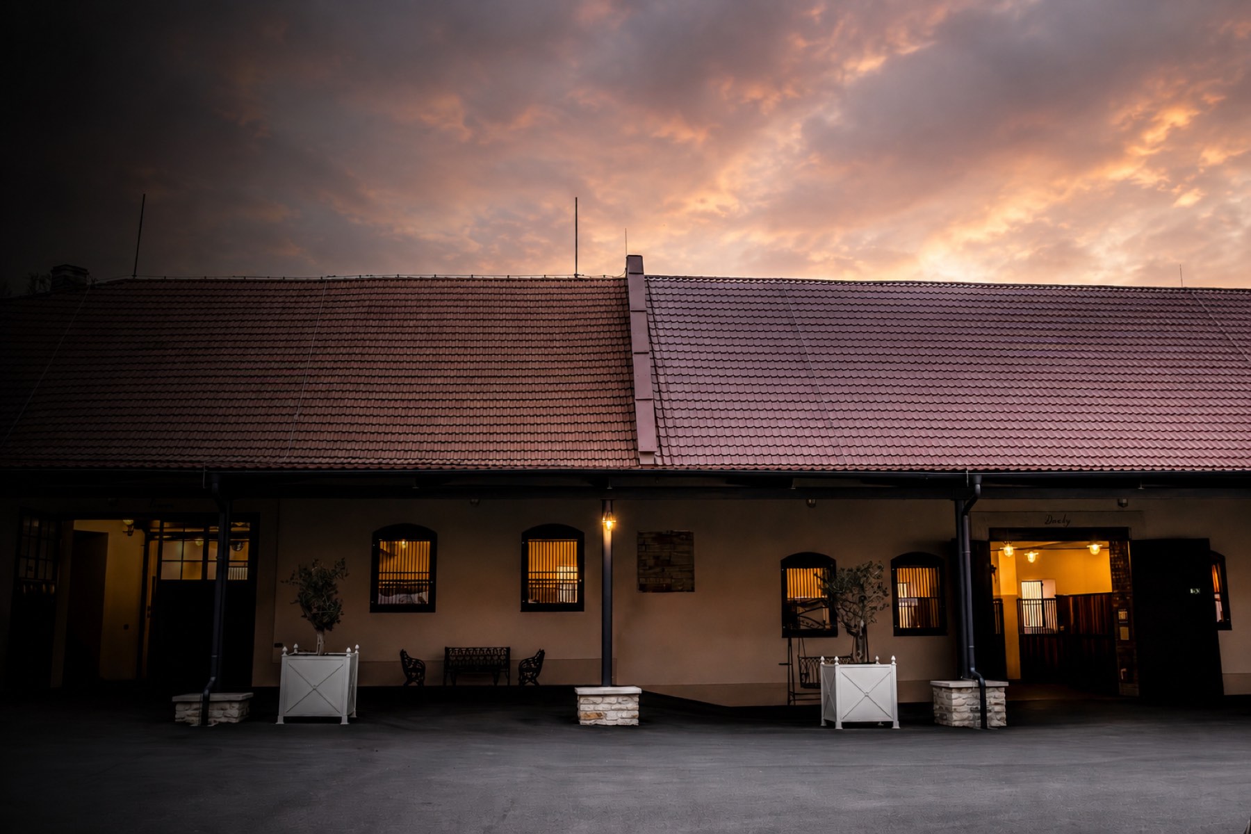 The estate at sunset, with lit windows, olive trees in planters, and a dramatic sky