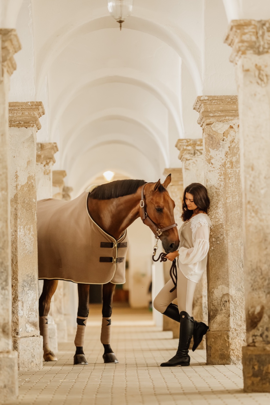 Emma Sophia Španko standing with her horse beneath the vaulted stone arches of the stable